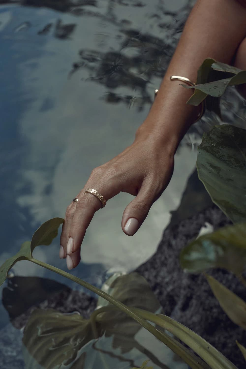 A hand wearing gold rings and a bangle, resting over still water