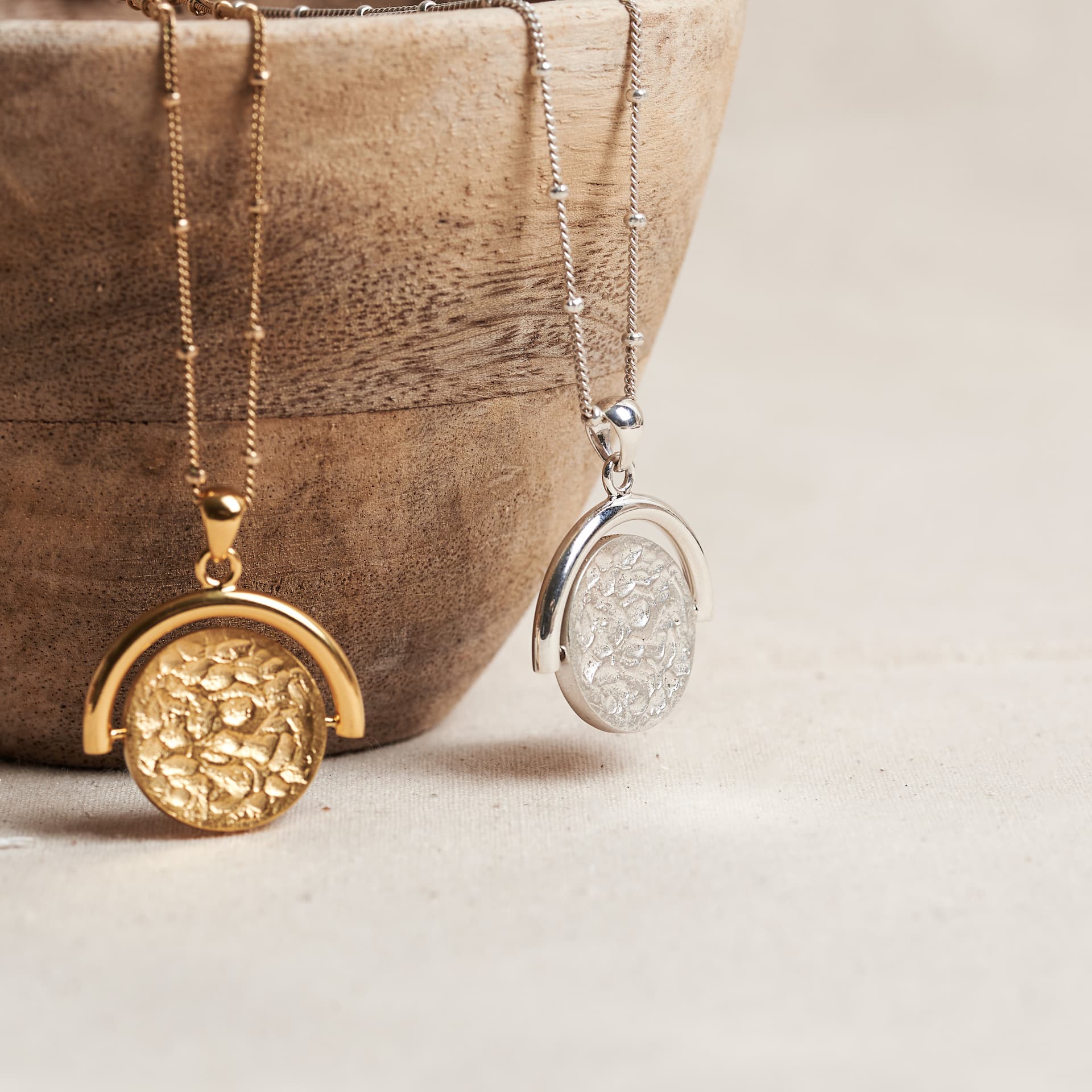 A hand-finished gold coin pendant and its silver twin, resting against a turned wooden bowl